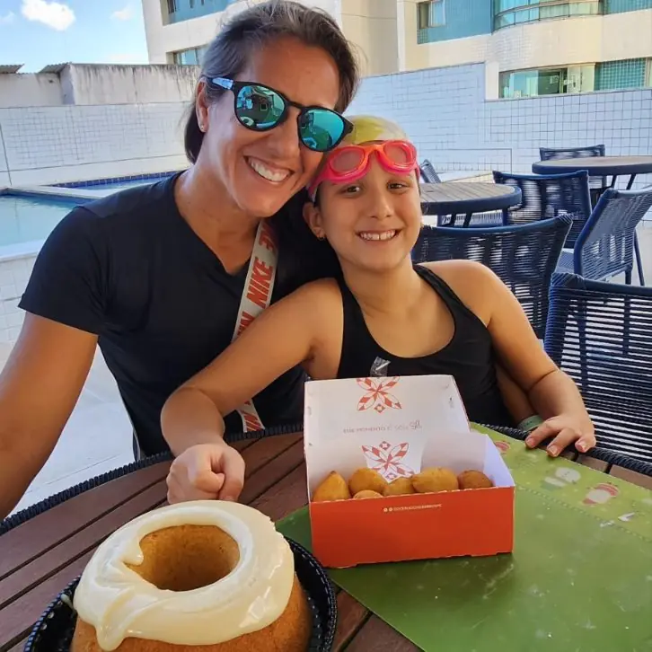 Natação infantil em casa: Roberta abraçada com uma aluna à beira da piscina do condomínio